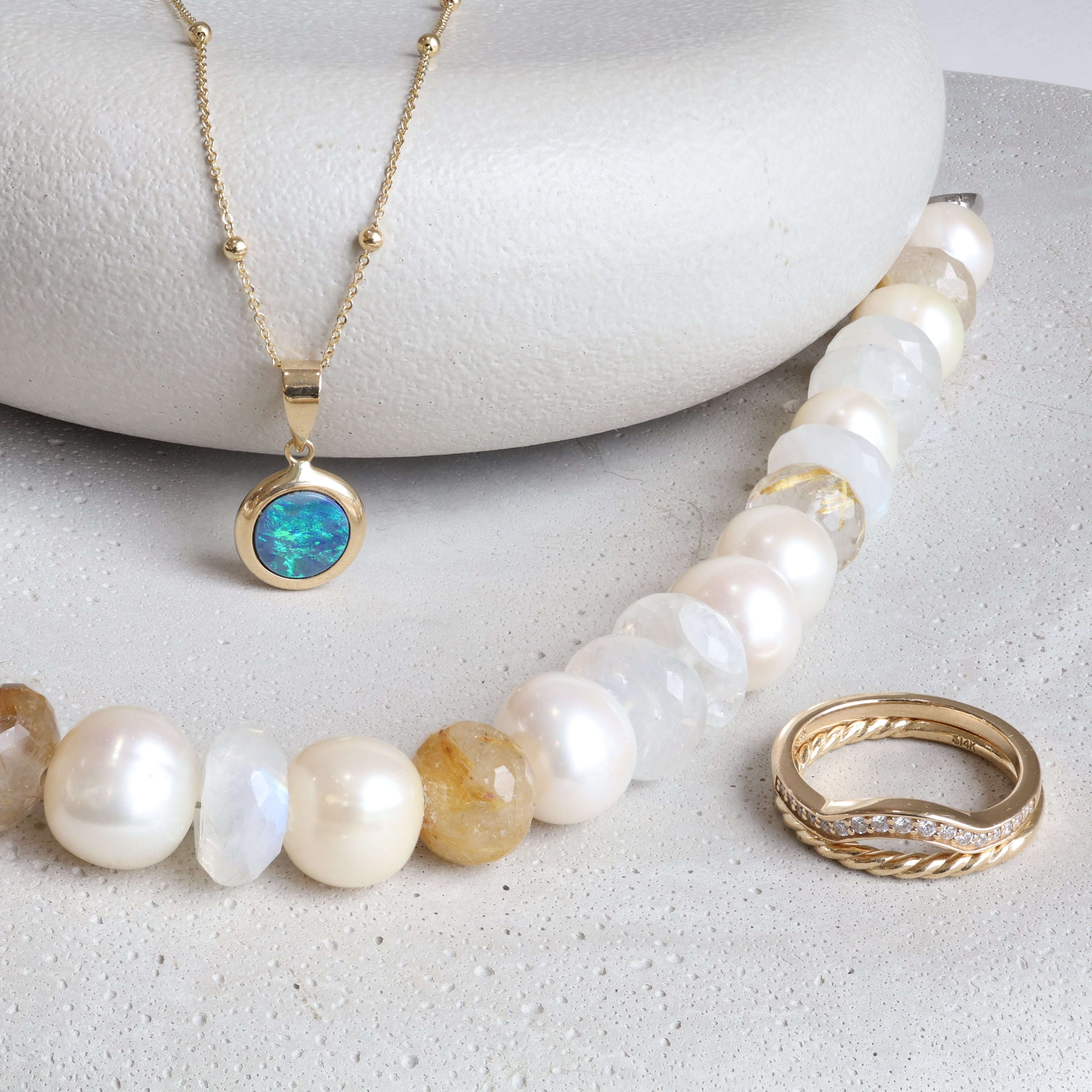 Opal and gold necklace, white beaded necklace, and a gold and diamond ring arranged on two stones.